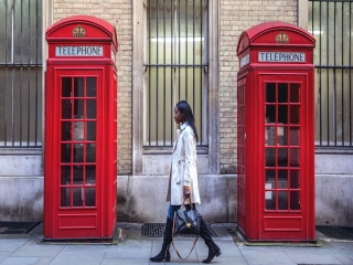 woman in trenchcoat walking down the street in london passing by two red phone booths