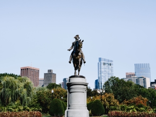 A Statue Of A Person Holding A Spear In A Park With Trees And Buildings In The Background